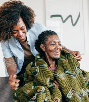 Caregiver wrapping a warm blanket around an older woman with dementia, smiling together.