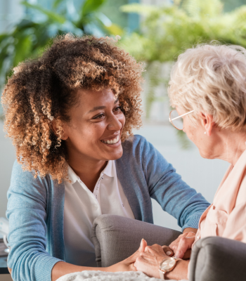 Caregiver smiling and holding hands with an older woman in a sunlit living room.
