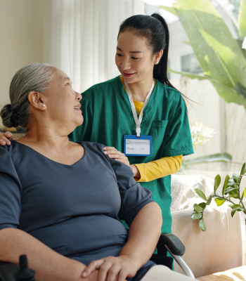 Caregiver in green scrubs leaning in to listen to an older woman in a wheelchair at home.