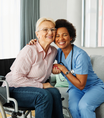 Smiling caregiver sitting beside an older woman in a wheelchair at home in Metro Detroit.